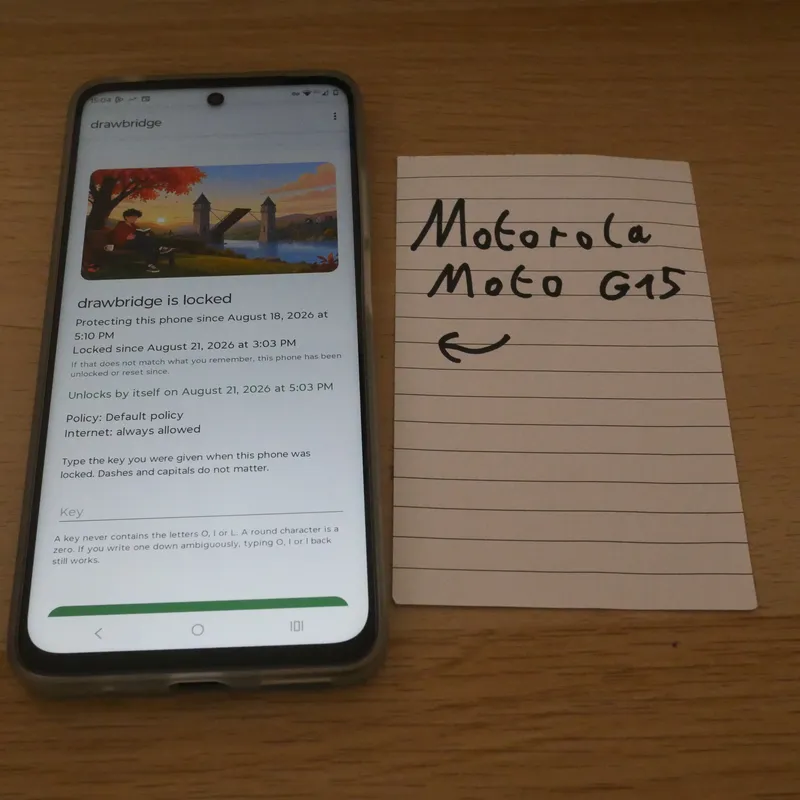 A Motorola Moto G15 on a table showing drawbridge's locked screen, beside a hand-written card naming the phone.