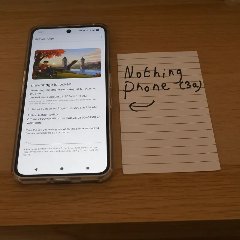 A Nothing Phone (3a) on a table showing drawbridge's locked screen with an evening curfew, beside a hand-written card naming the phone.